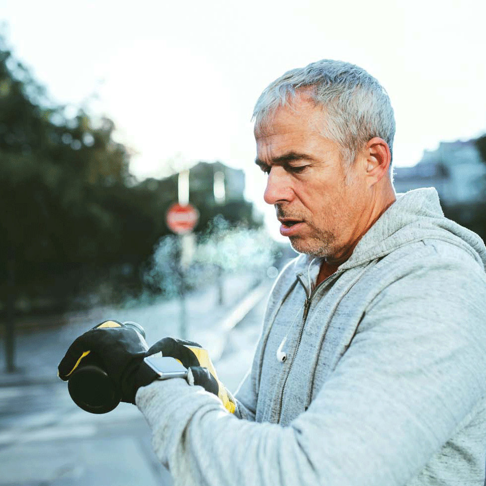 close-up of a middle aged man wearing a grey sports jumper and black gloves, looking at his smart watch, blurred city background 