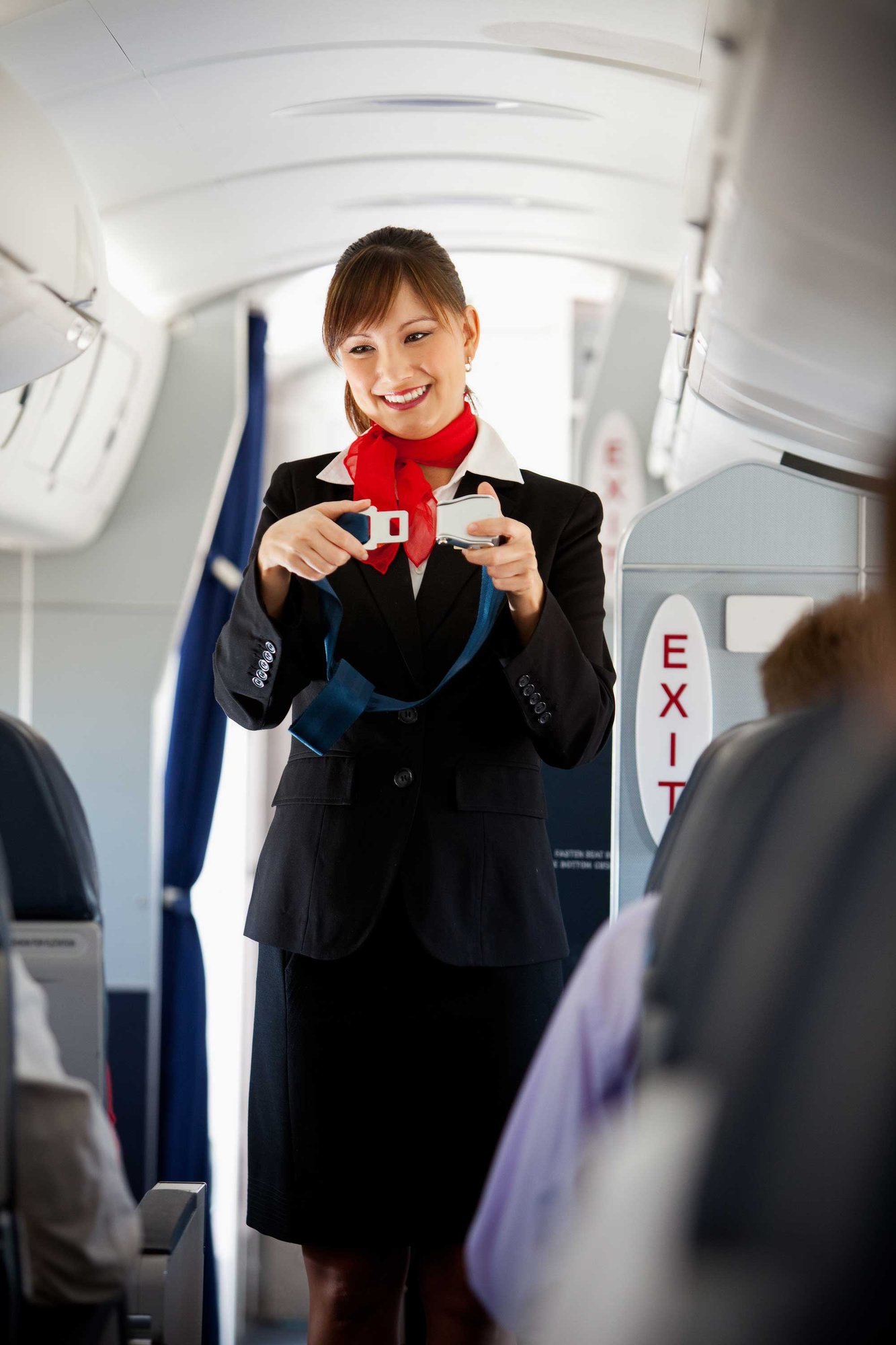 A female flight attendant demonstrating safety procedures to passengers, she is smiling, has a seatbelt in her hand