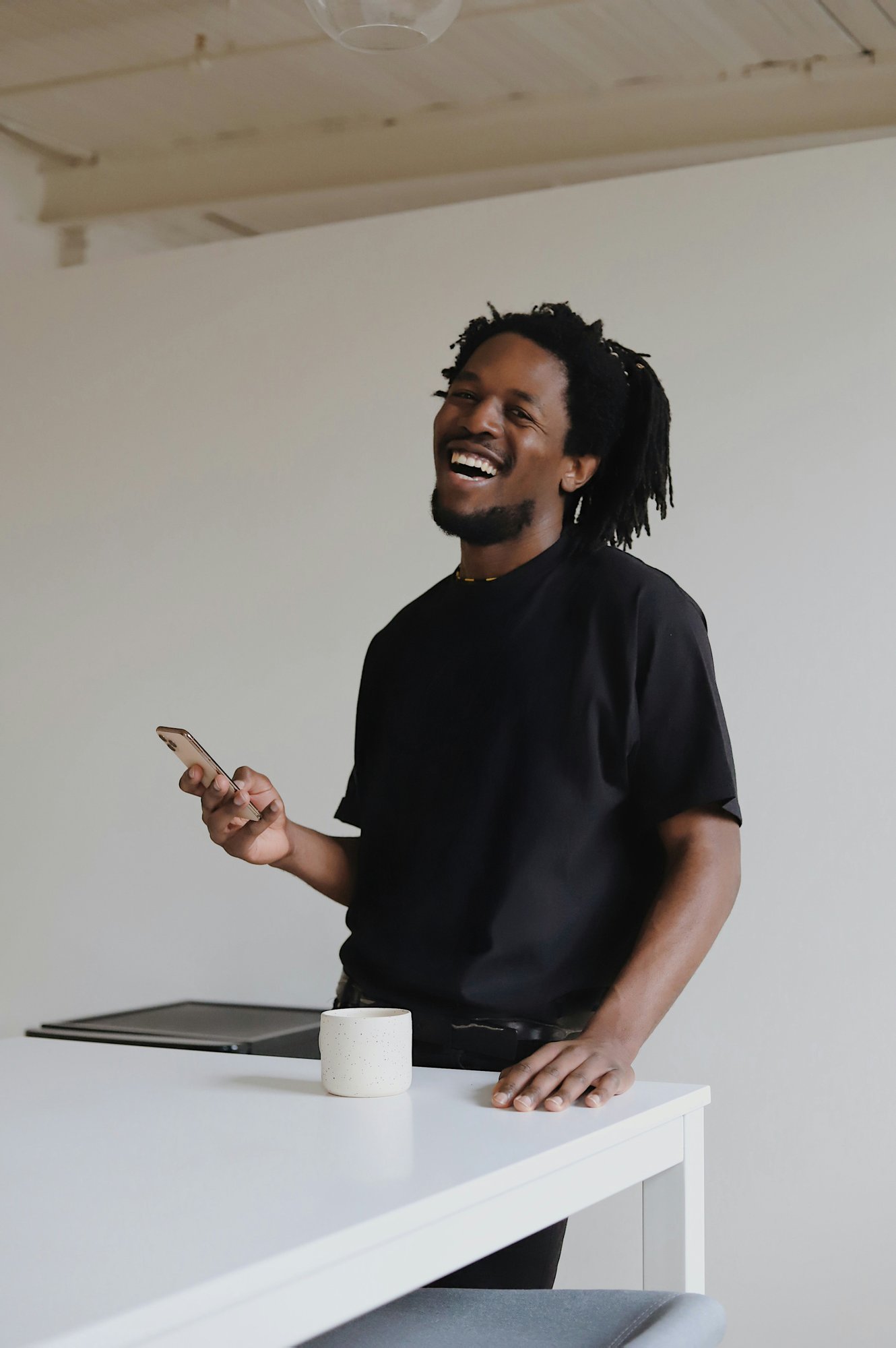 a black man standing behind a white table, smiling brightly, holding a mobile phone, on the table are a closed laptop and a white cup