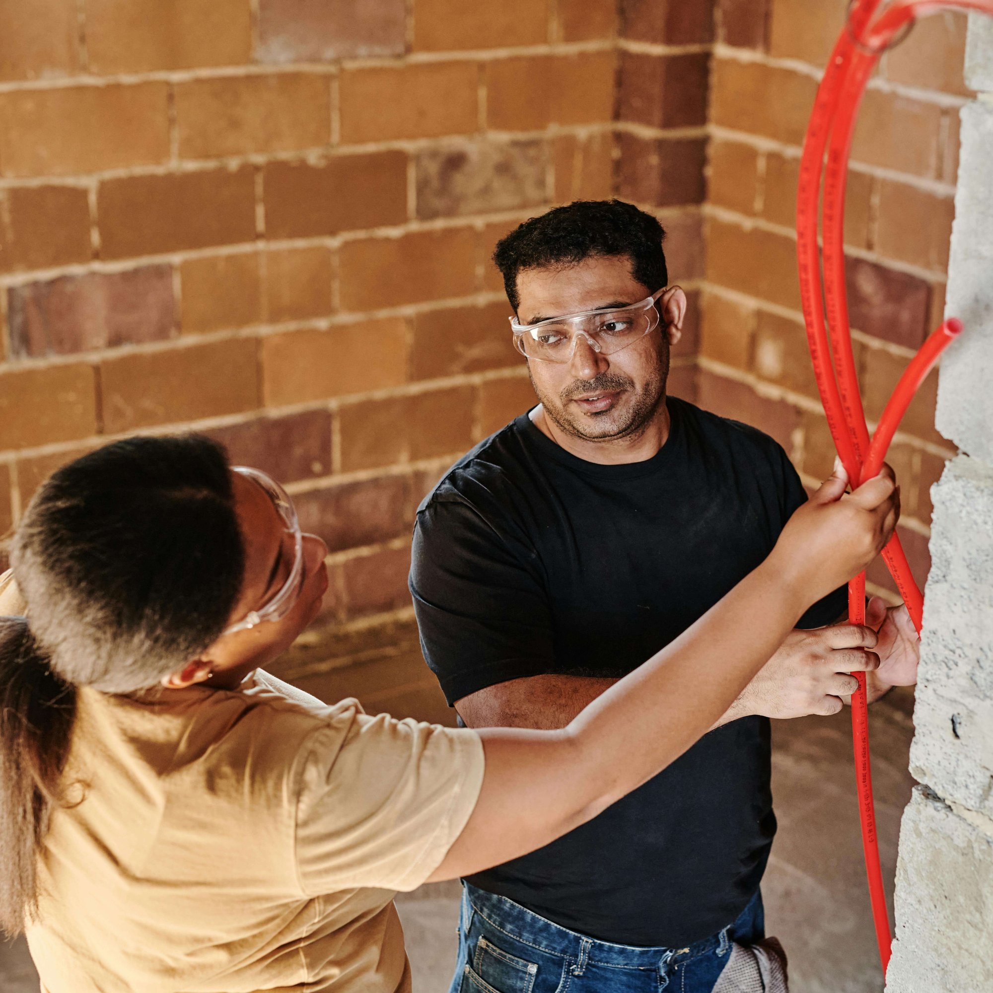 a man and a woman on a building site, in front of a brick wall, looking at a  some red pipes, in conversation, wearing safety googles